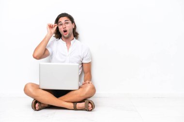 Young handsome man with a laptop sitting on the floor isolated on white background with glasses and surprised