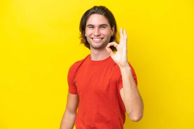 Sarı arka planda izole edilmiş beyaz yakışıklı adam parmaklarıyla tamam işareti gösteriyor.