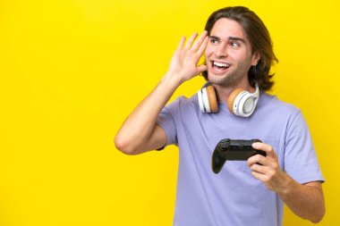 Genç, beyaz bir adam, sarı arka planda izole edilmiş bir bilgisayar oyunu kontrolörüyle oynuyor. Kulağına elini sürerek bir şeyler dinliyor.