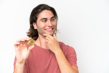 Beyaz arka planda izole edilmiş Bitcoin tutan genç yakışıklı bir adam bir fikir ve bir bakış açısı düşünüyor.