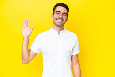 Genç yakışıklı adam izole edilmiş sarı arka planda mutlu bir ifadeyle selamlıyor.