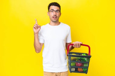 Elinde yiyecek dolu bir alışveriş sepeti olan genç yakışıklı bir adam izole edilmiş sarı arka planda parmağını kaldıran bir fikir düşünüyor.