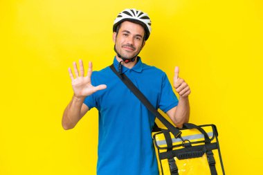 Termal sırt çantalı yakışıklı genç adam izole edilmiş sarı arka planda altı parmaklı sayıyor.