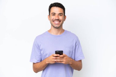 İzole edilmiş beyaz arka planda genç yakışıklı adam kameraya bakıyor ve cep telefonunu kullanırken gülümsüyor.