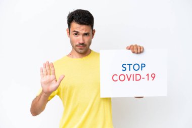Beyaz arka plan üzerinde genç yakışıklı bir adam elinde yazılı bir levha tutuyor. Dur Covid 19 ve dur işareti yapıyor.