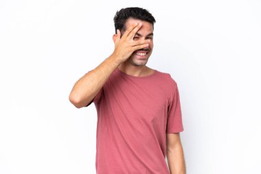 İzole edilmiş beyaz arka planda genç yakışıklı bir adam. Gözleri elleriyle kaplıyor ve gülümsüyor.