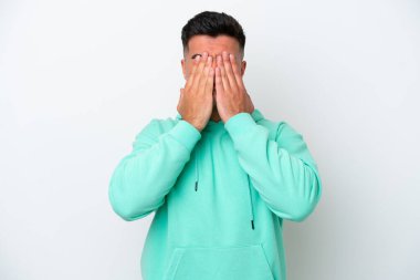 Beyaz arka planda izole edilmiş beyaz tenli yakışıklı adam gözlerini elleriyle kapatıyor.