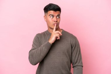 Pembe arka planda izole edilmiş beyaz, yakışıklı genç bir adam sessiz bir jest işareti gösteriyor. Parmağını ağzına sokuyor.