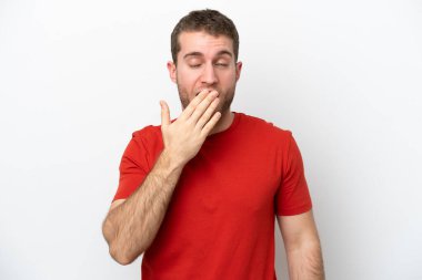 Beyaz arka planda izole edilmiş genç beyaz bir adam esniyor ve geniş bir ağzı eliyle kapatıyor.