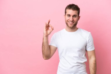 Pembe arka planda izole edilmiş genç beyaz adam parmaklarıyla tamam işareti gösteriyor.