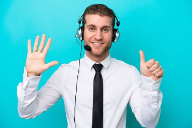Tele pazarlamacı beyaz adam mavi arka planda izole edilmiş bir kulaklıkla çalışıyor. Parmakları olan altı kişi sayıyor.