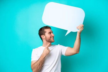 Young caucasian man isolated on blue background holding an empty speech bubble and pointing it