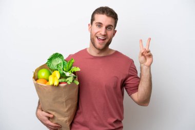 Young caucasian man holding a grocery shopping bag isolated on white background smiling and showing victory sign