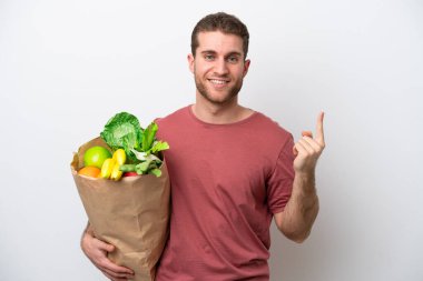 Beyaz arka planda izole edilmiş alışveriş torbası tutan genç beyaz bir adam en iyinin işaretini gösteriyor ve parmağını kaldırıyor.
