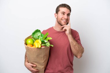 Beyaz arka planda izole edilmiş alışveriş torbası tutan genç beyaz bir adam yukarı bakarken bir fikir düşünüyor.