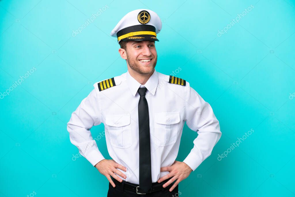 Airplane caucasian pilot isolated on blue background posing with arms at hip and smiling