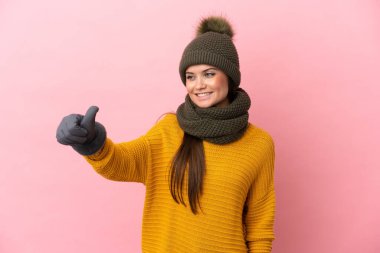 Pembe arka planda izole edilmiş beyaz şapkalı genç bir kız baş parmak hareketi yapıyor.