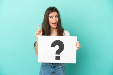 Kafkasyalı genç bir kadın mavi arka planda izole edilmiş. Elinde soru işareti olan bir levha var ve düşünüyor.
