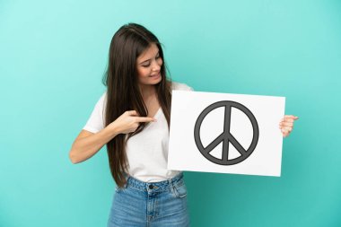 Kafkasyalı genç bir kadın mavi arka planda izole edilmiş. Elinde barış sembolü olan bir levha var.