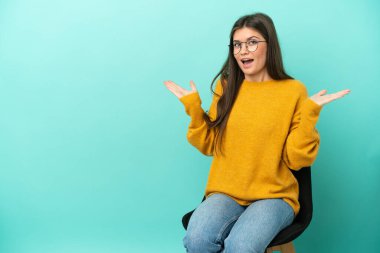 Mavi arka planda izole edilmiş bir sandalyede oturan beyaz kadın, şok olmuş yüz ifadesiyle.