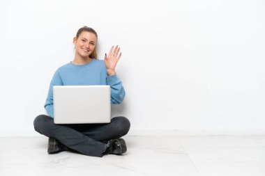 Dizüstü bilgisayarı olan genç bir kadın yerde oturmuş mutlu bir ifadeyle selam veriyor.