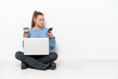 Dizüstü bilgisayarı olan genç bir kadın elinde kahve ve cep telefonu ile yerde oturuyor.