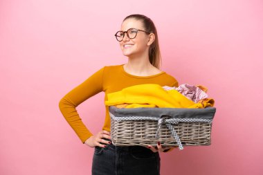 Pembe arka planda, kolları kalçasında poz veren ve gülümseyen bir çamaşır sepetini tutan beyaz bir kadın.