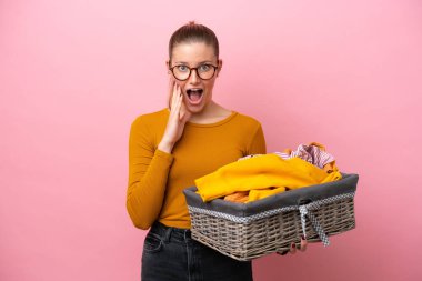 Pembe arka planda yüzü şoke olmuş ve şaşırmış bir halde kıyafet sepeti tutan beyaz bir kadın.
