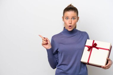Young caucasian woman holding a gift isolated on white background surprised and pointing side