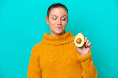 Avokado tutan beyaz bir kadın mavi arka planda mutlu bir ifadeyle izole edilmiş.