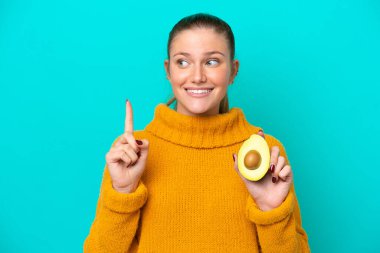 Avokado tutan beyaz bir kadın mavi arka planda izole edilmiş. Çözümü bulmak için parmağını kaldırıyor.