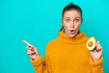 Avokado tutan beyaz bir kadın mavi arka planda izole edilmiş. Şaşırmış ve işaret ediyor.