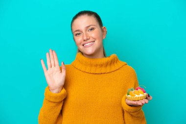 Mavi arka planda izole edilmiş bir tart tutan beyaz bir kadın mutlu bir ifadeyle selamlıyor.
