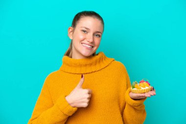 Kafkasyalı genç bir kadın mavi arka planda izole edilmiş bir tart tutuyor. Başparmakları havada çünkü iyi bir şey oldu.