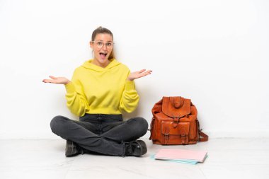 Beyaz arka planda beyaz bir zeminde oturan beyaz bir kadın, şok olmuş bir yüz ifadesiyle.