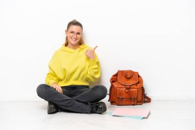 Beyaz arka planda beyaz bir zeminde oturan beyaz tenli genç bir kadın bir ürünü sunmak için yan tarafı işaret ediyor.