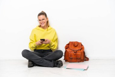 Beyaz arka planda beyaz bir zeminde oturan genç beyaz kadın cep telefonuyla bir mesaj gönderiyor.