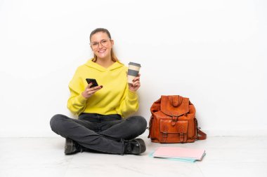 Beyaz arka planda bir katta oturan beyaz tenli genç bir kadın kahve alıp götürmek için elinde bir cep telefonu var.