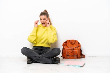 Beyaz arka planda beyaz bir zeminde oturan beyaz tenli genç bir kadın gözlüklü ve şaşırmış.