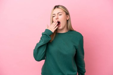 Pembe arka planda izole edilmiş genç beyaz bir kadın esniyor ve geniş bir ağzı eliyle kapatıyor.