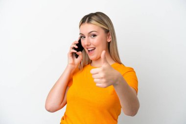 Beyaz arka planda izole edilmiş genç beyaz kadın başparmaklarını kaldırırken cep telefonuyla sohbet ediyor.