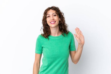 Kıvırcık saçlı genç bir kadın beyaz arka planda izole edilmiş mutlu bir ifadeyle selam veriyor.