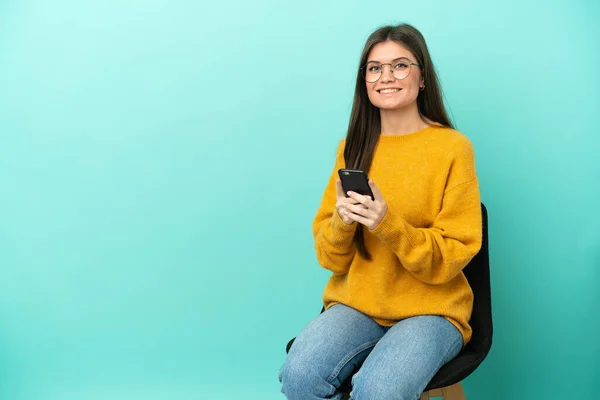 Young caucasian woman sitting on a chair isolated on blue background sending a message with the mobile