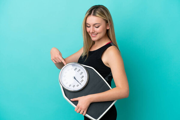 Young caucasian woman isolated on blue background holding a weighing machine and pointing it
