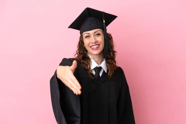 Young university graduate woman isolated on pink background shaking hands for closing a good deal
