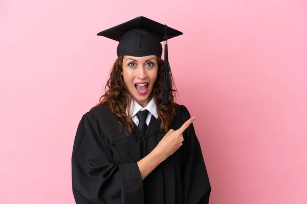 Young university graduate woman isolated on pink background surprised and pointing side
