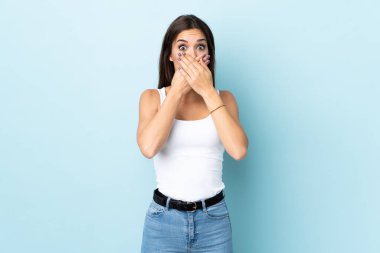 Mavi arka planda izole edilmiş genç beyaz kadın elleriyle ağzını kapatıyor.