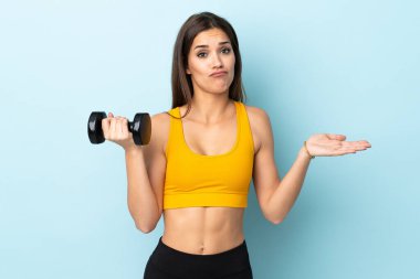 Beyaz bir kadın halter kaldırıyor. Mavi arka planda tek başına. Omuzlarını kaldırırken şüphe uyandırıyor.