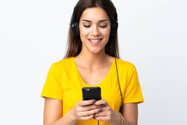 Beyaz arka planda izole edilmiş genç beyaz kadın müzik dinliyor ve cep telefonu arıyor.
