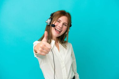 Tele pazarlamacı İngiliz kadın mavi arka planda izole edilmiş bir kulaklıkla çalışıyor. Başparmakları havada çünkü iyi bir şey oldu.